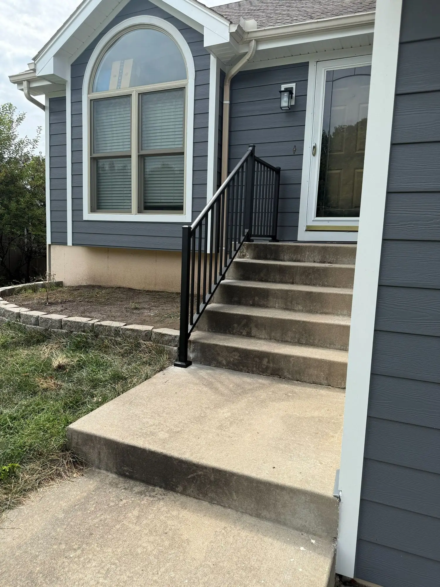 Concrete front steps and walkway with a new black metal handrail against grey-blue siding